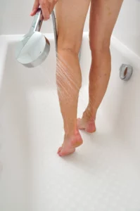 Eine Person steht in einer Dusche und hält einen Handbrausekopf, der Wasser auf ihre Unterschenkel und Füße sprüht.