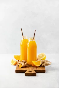 Zwei Flaschen mit frischem Orangensaft und Strohhalmen auf einem Holzbrett, umgeben von Orangenschnitzen und einem Stück Ingwer, vor einem hellen Hintergrund.