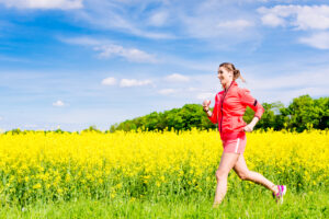 Eine fröhliche Läuferin mit Sportkleidung und Ohrhörern joggt durch ein blühendes Rapsfeld an einem sonnigen Tag. Der Himmel ist klar und blau mit einigen lockeren Wolken. Sie scheint die Bewegung und die frische Luft zu genießen, während die Natur um sie herum in sattem Gelb erstrahlt.